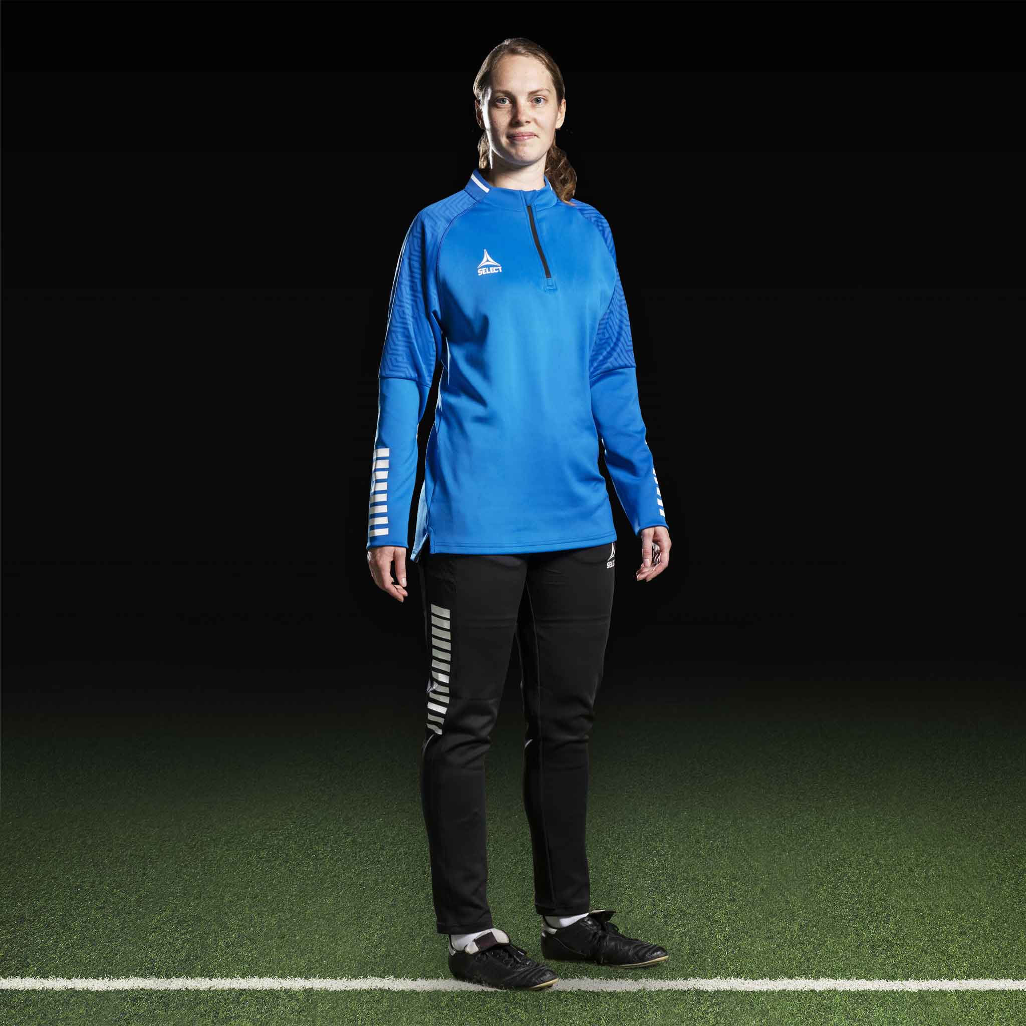 Kvindelig fodboldspiller i blå Training sweat 1/2 zip Monaco v24, Match & Training wear, med sorte bukser, set forfra.
