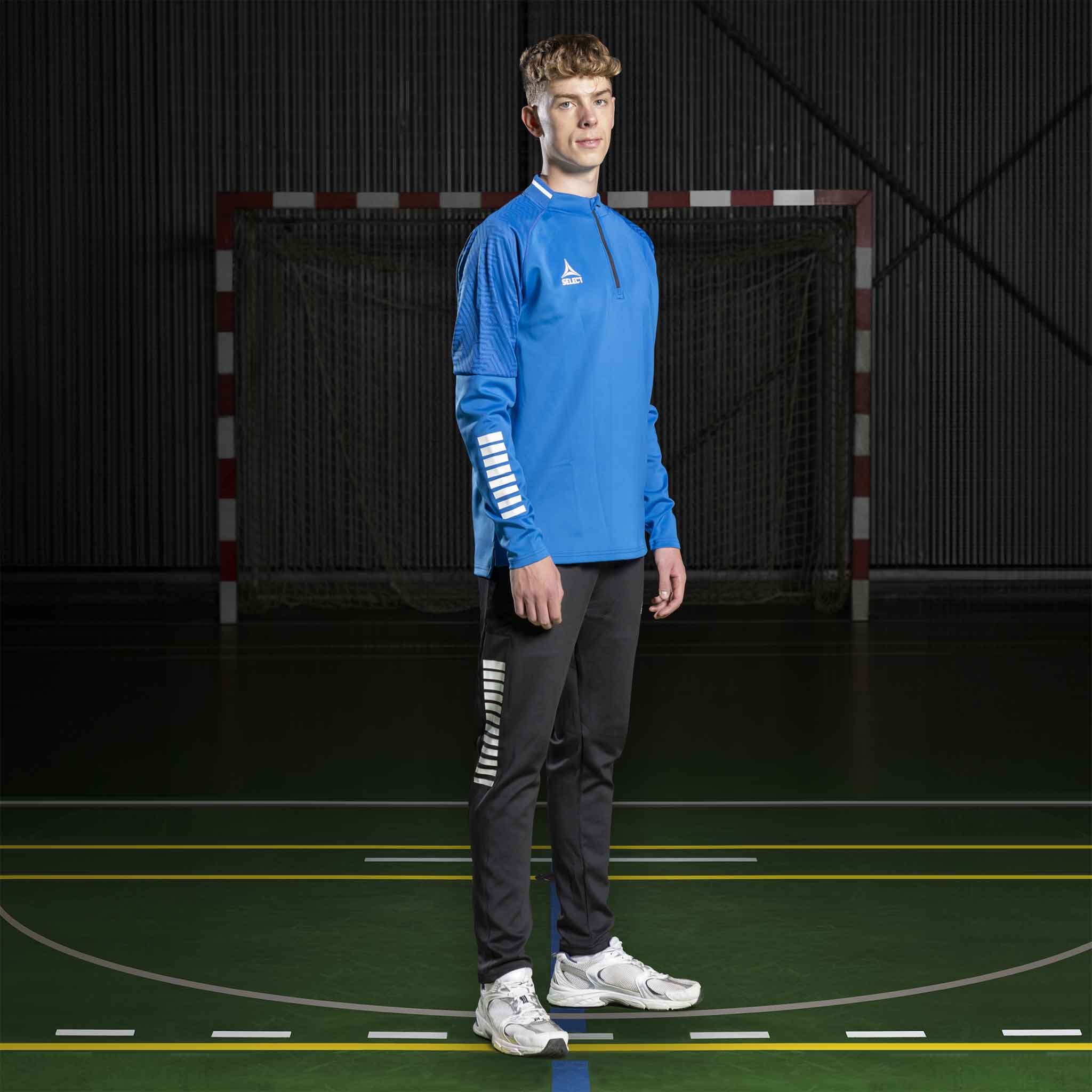 Mandlig håndboldspiller i blå Training sweat 1/2 zip Monaco v24 og sorte bukser, Match & Training wear, forfra.