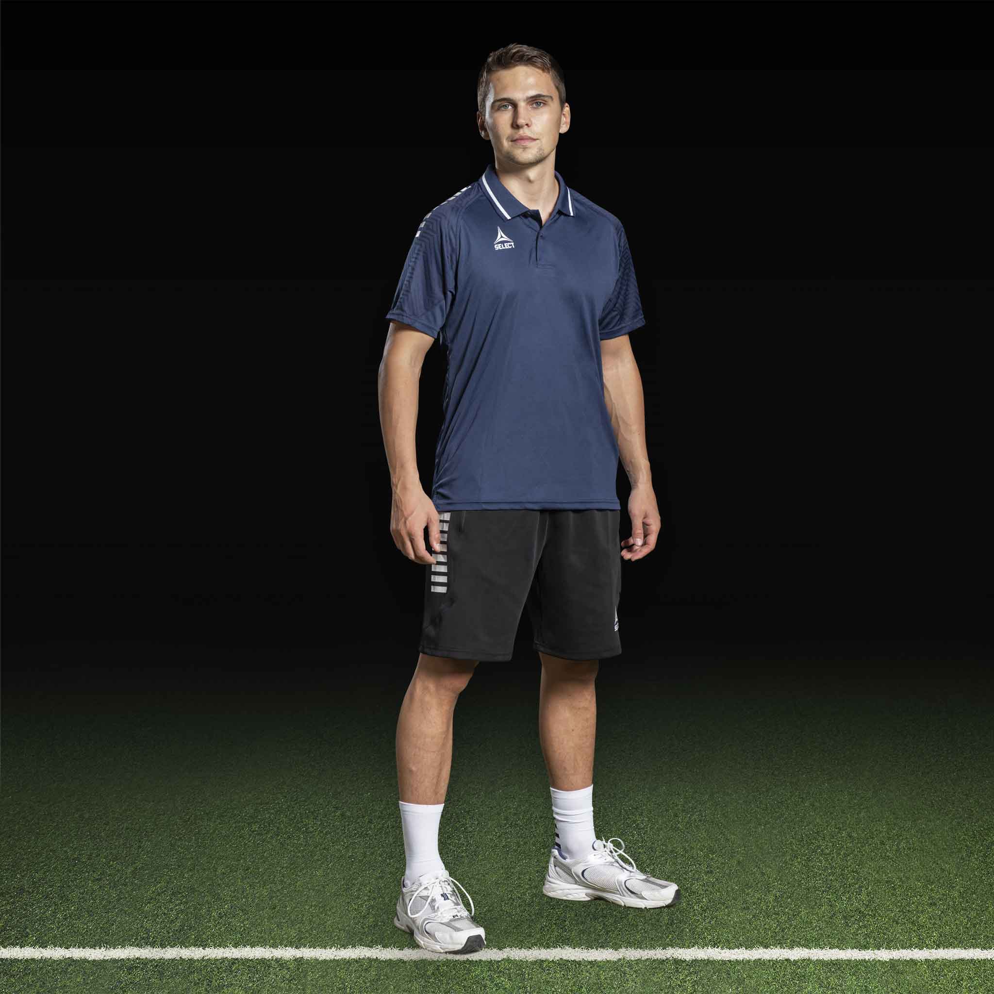 Mandlig fodboldspiller i marineblå polo og sorte Bermuda shorts Monaco v24, leisure wear, forfra på banen.