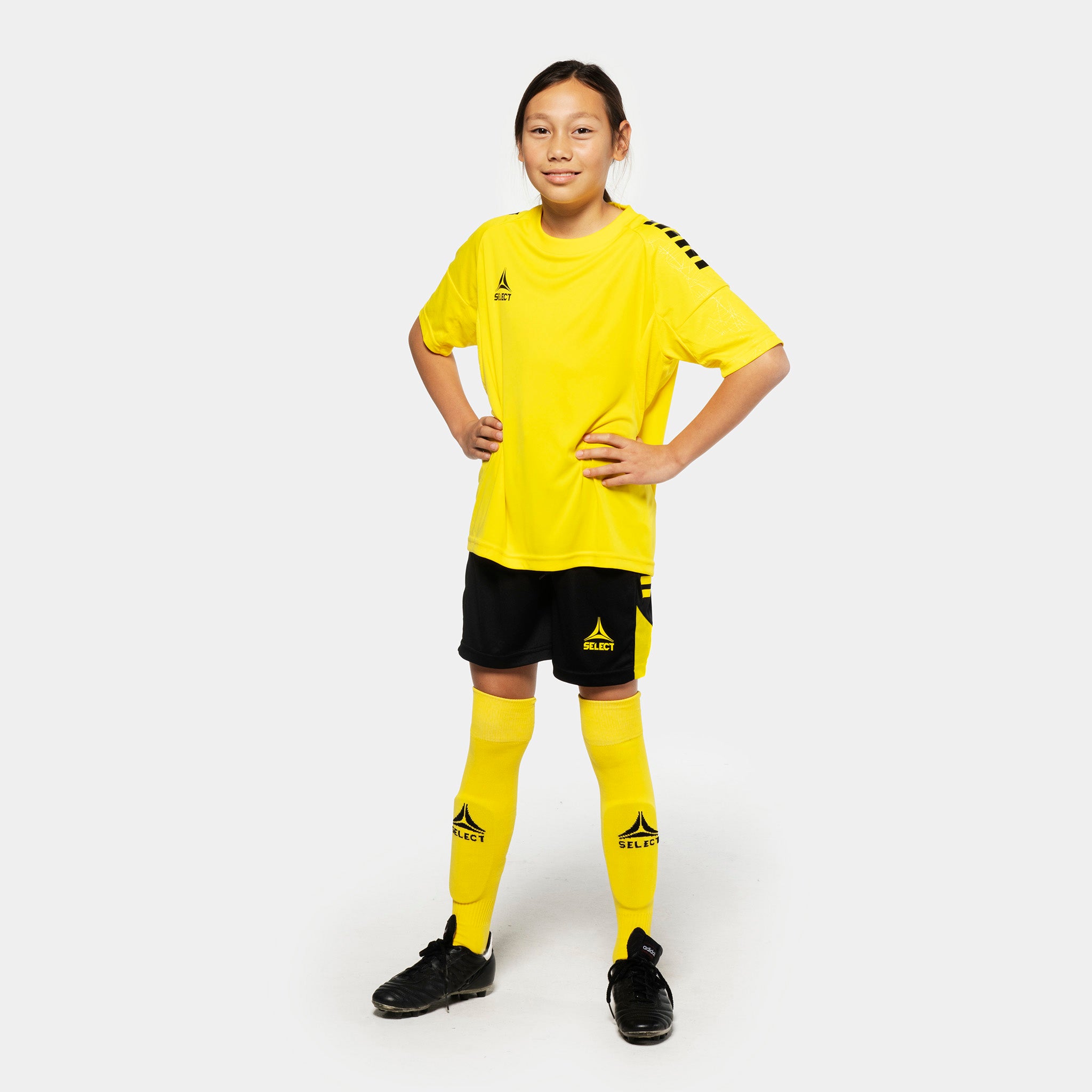 Pige fodboldspiller i gul trøje, sorte Player shorts Spain v25 og gule strømper. Match & Training wear, forfra.