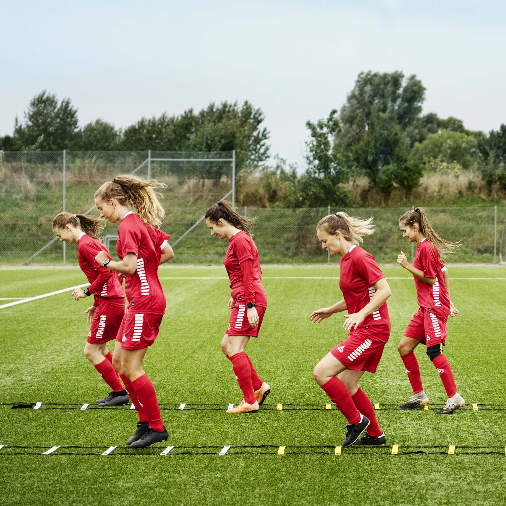Kvindelige fodboldspillere træner med orange og gul Agility ladder, fodbold træningsudstyr, på grøn bane.